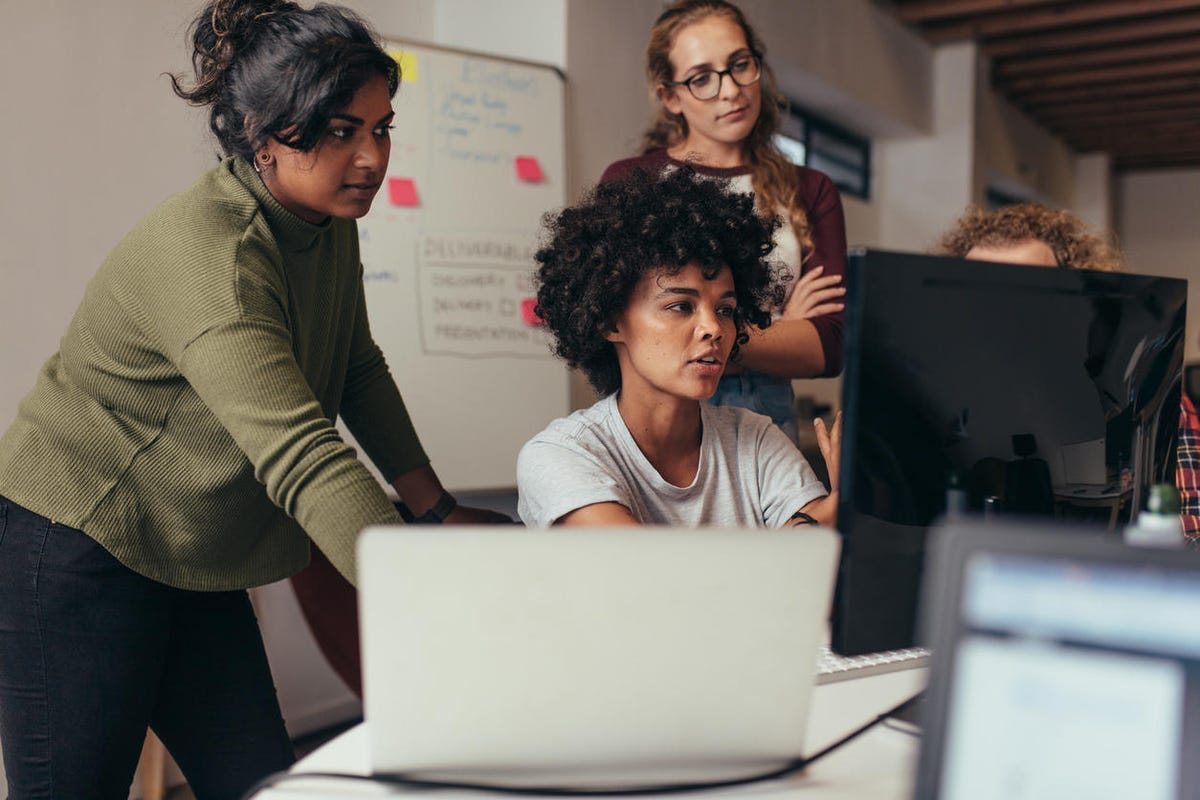 Wanita muda multietnis bekerja bersama sambil berdiri dan duduk di depan komputer